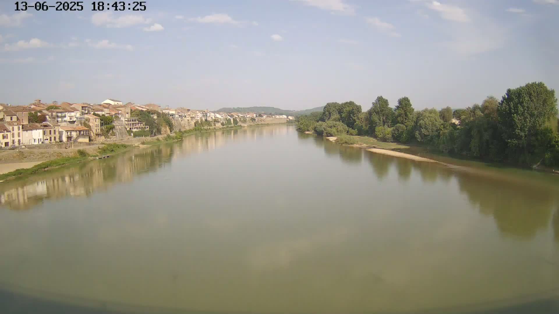 A wide, calm river reflects a town of buildings and trees lining one bank under a partly cloudy sky.