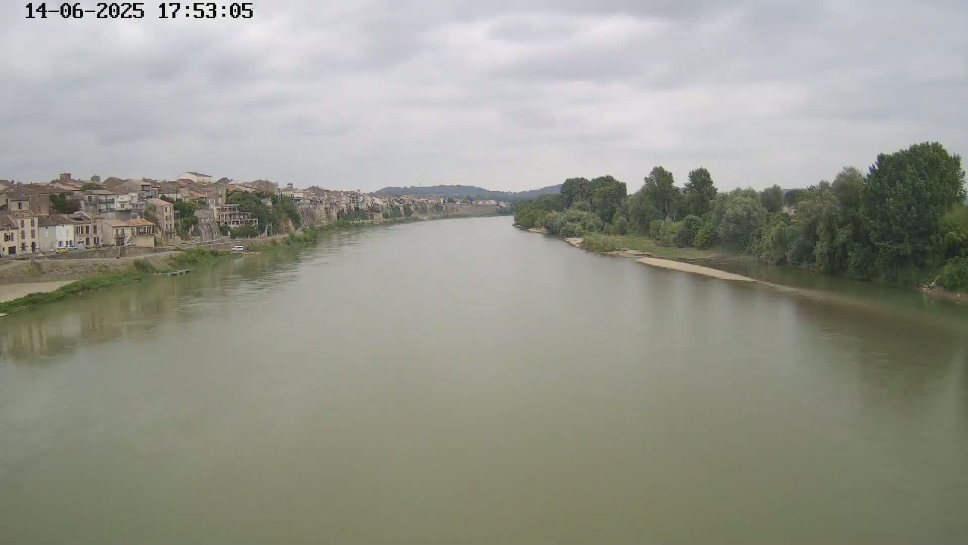A wide, muddy river flows under an overcast sky past a town of old, stone buildings on one bank and lush green trees on the other.
