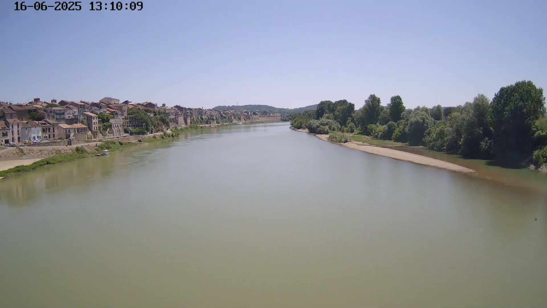 A wide, muddy river flows under a clear blue sky past a town on one bank and a line of trees on the other.