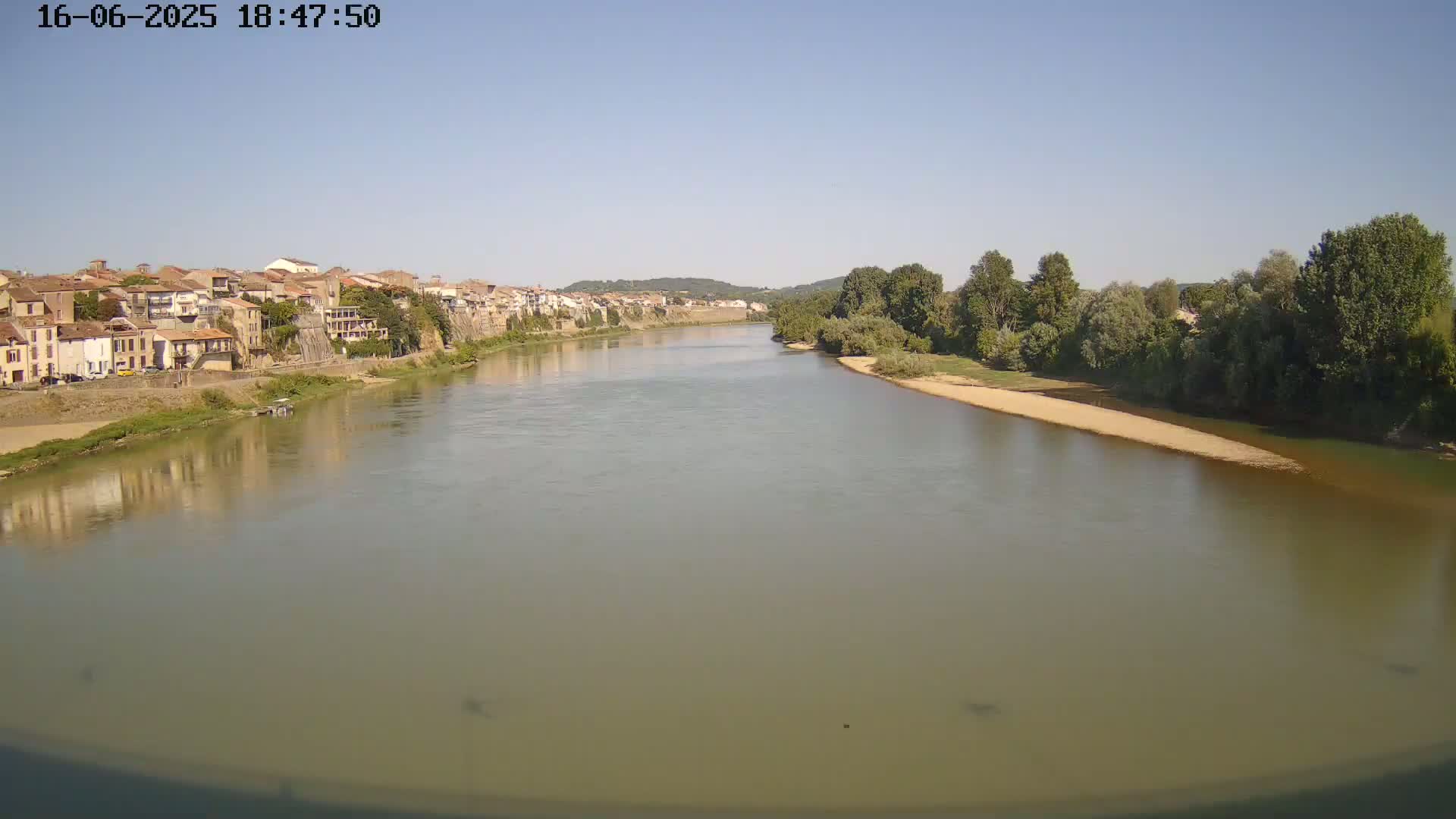 A wide, calm river flows past a town of buildings nestled against a hillside under a clear, sunny sky.