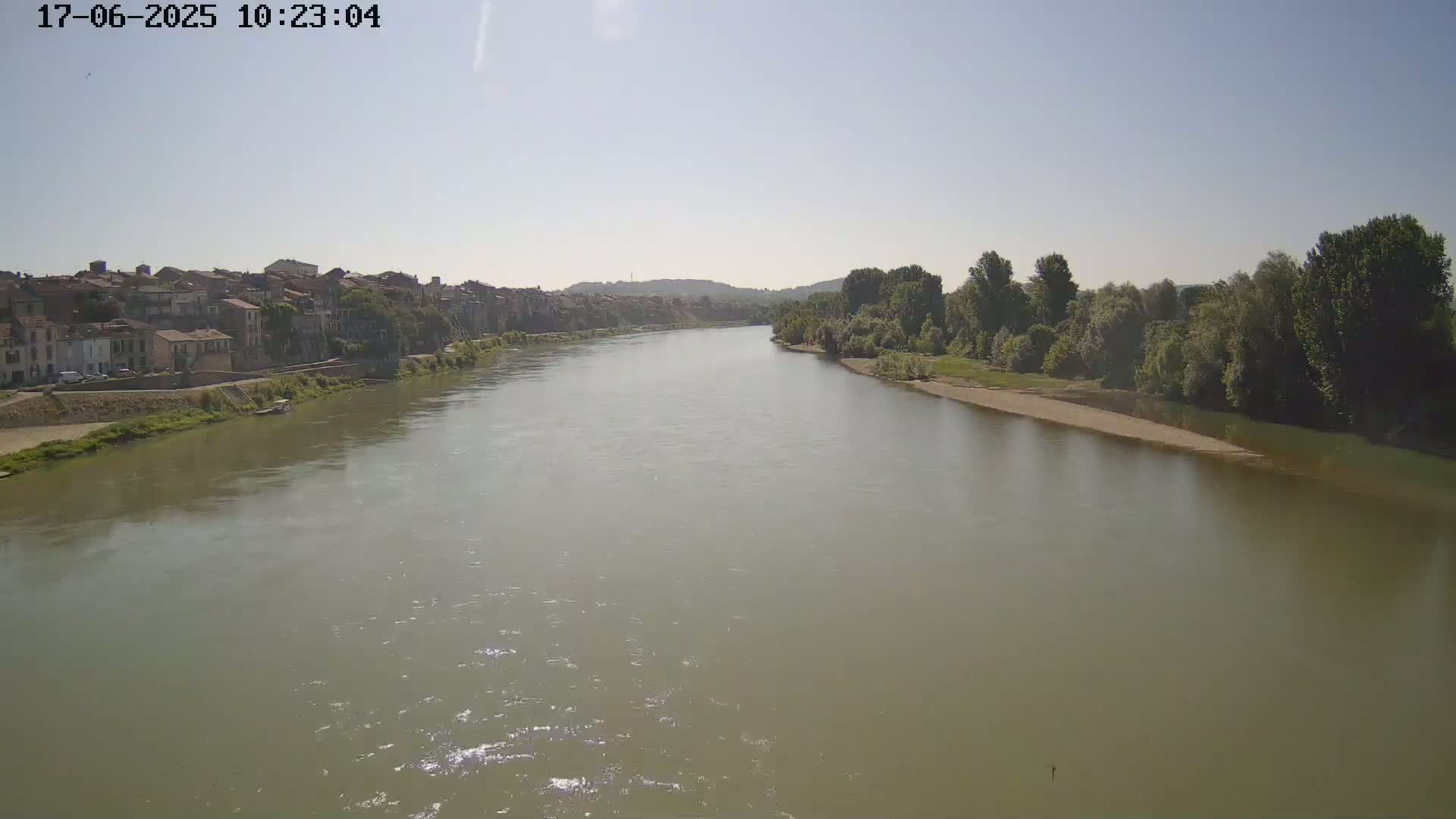 A wide, murky river flows past a town and lush green trees under a clear, sunny sky.