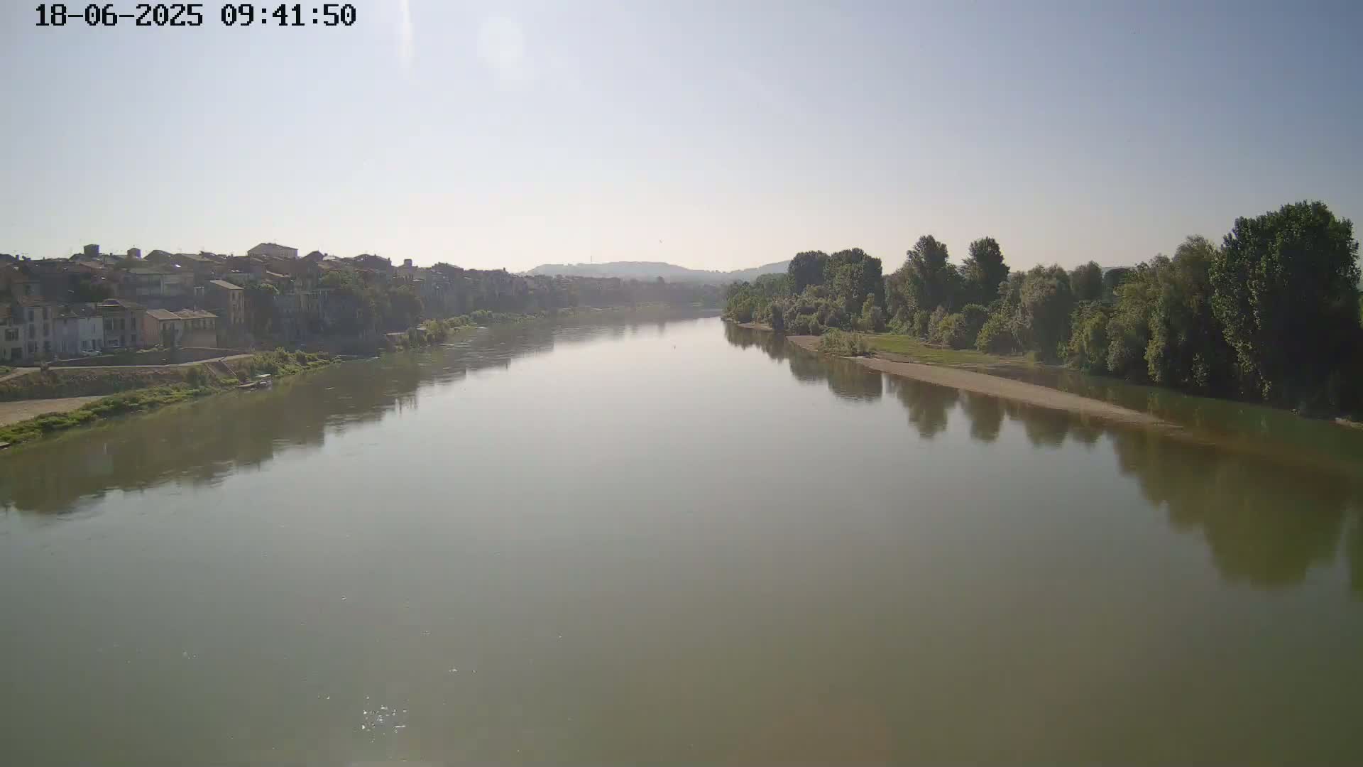A wide, calm river reflects the hazy sunlight and the buildings and trees along its banks under a clear sky.