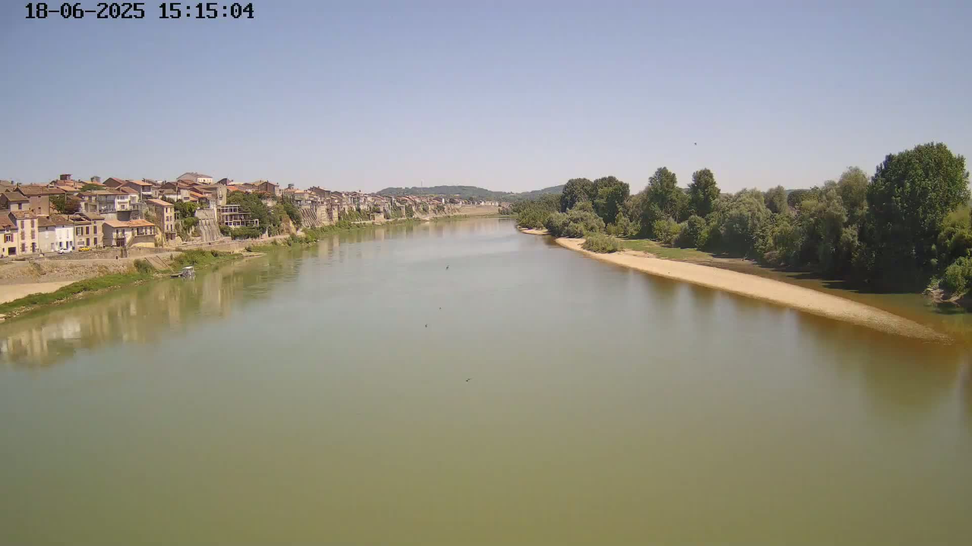 A wide, calm river flows past a town of old buildings on one bank and a sandy shore lined with trees on the other, under a clear sky.