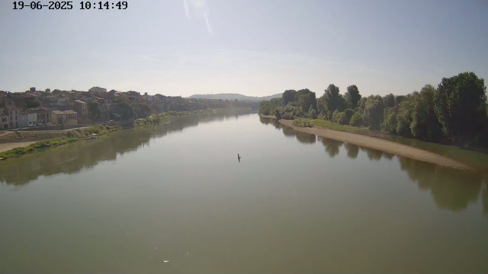 A wide, calm river reflects a mostly clear sky and the buildings and trees lining its banks.