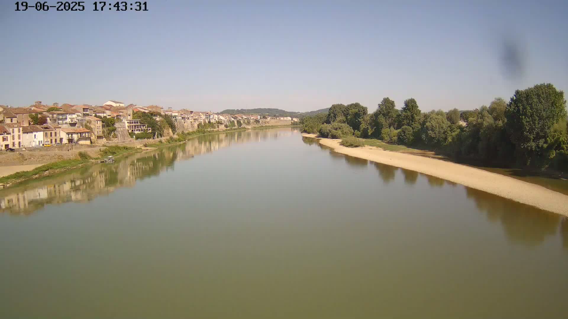 A calm river reflects a town of tan buildings on one bank and green trees on the opposite, sandy bank under a clear, sunny sky.