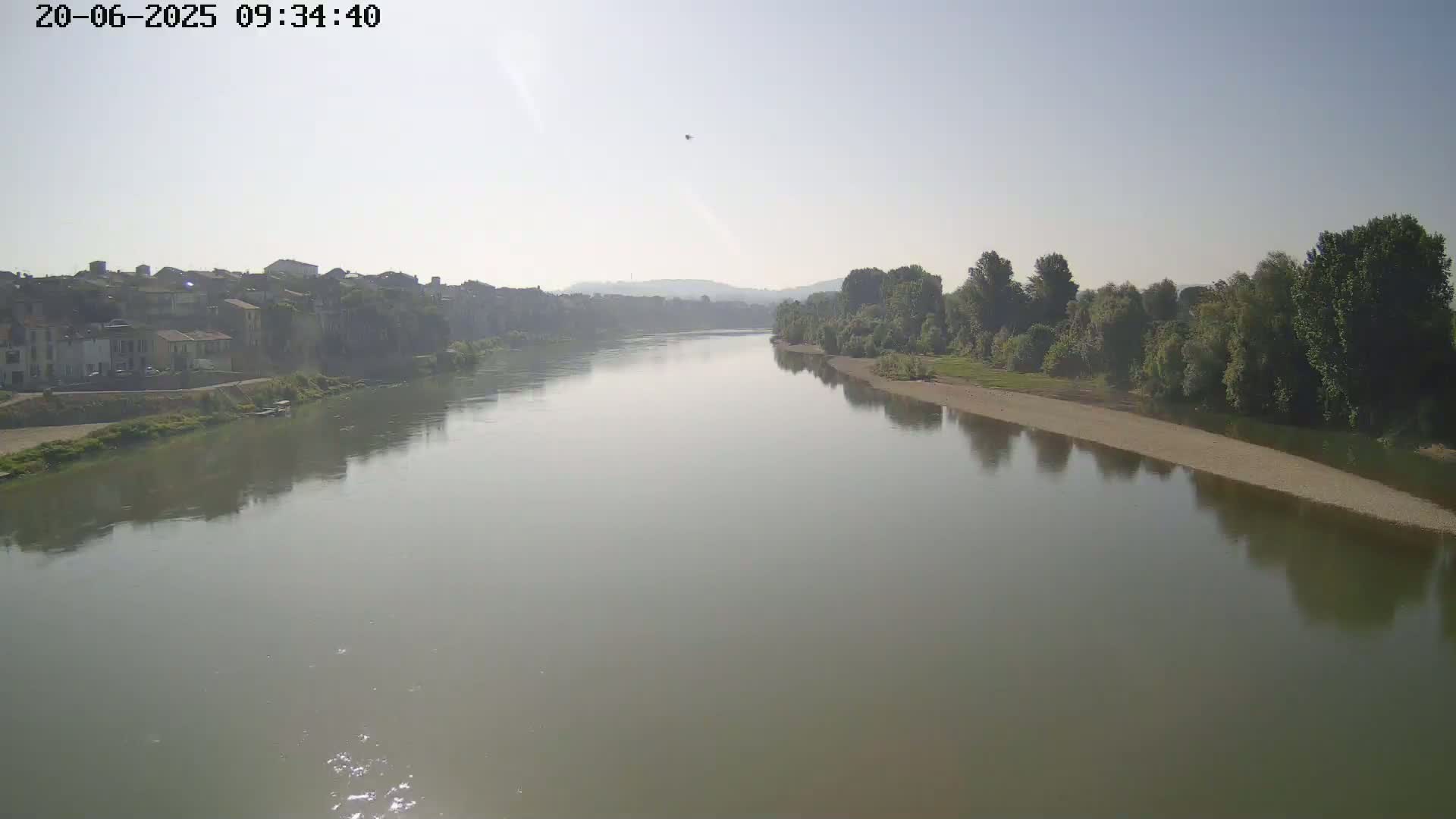 A wide, calm river reflects the hazy sky and a line of trees and buildings on its banks under a mostly clear sky.