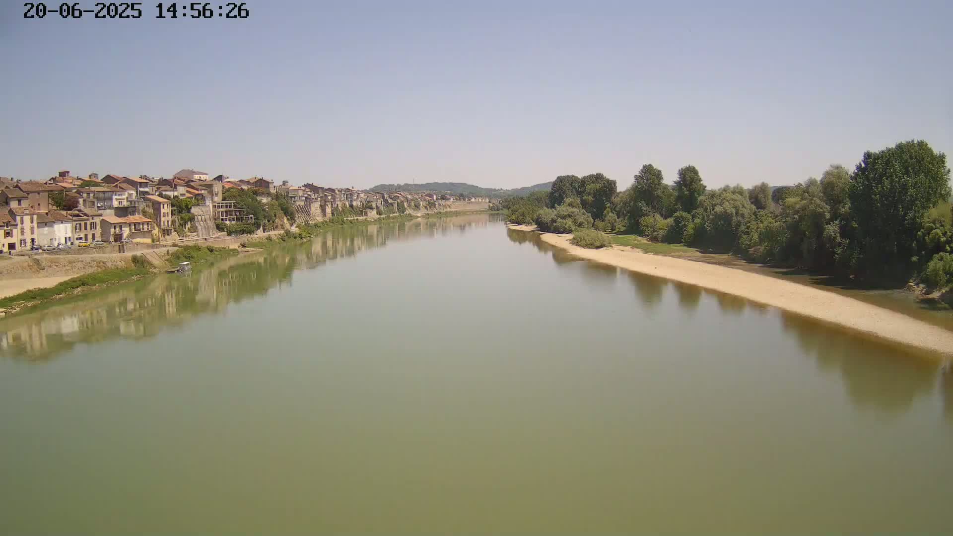 A wide, calm river reflects a town of buildings on one bank and trees along the other under a clear, bright sky.