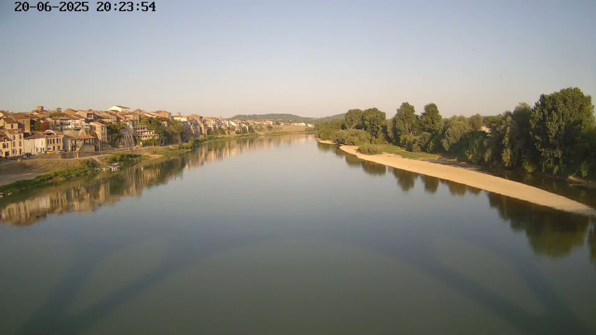 A calm river reflects a town of tan buildings and green trees under a clear, light-blue sky.