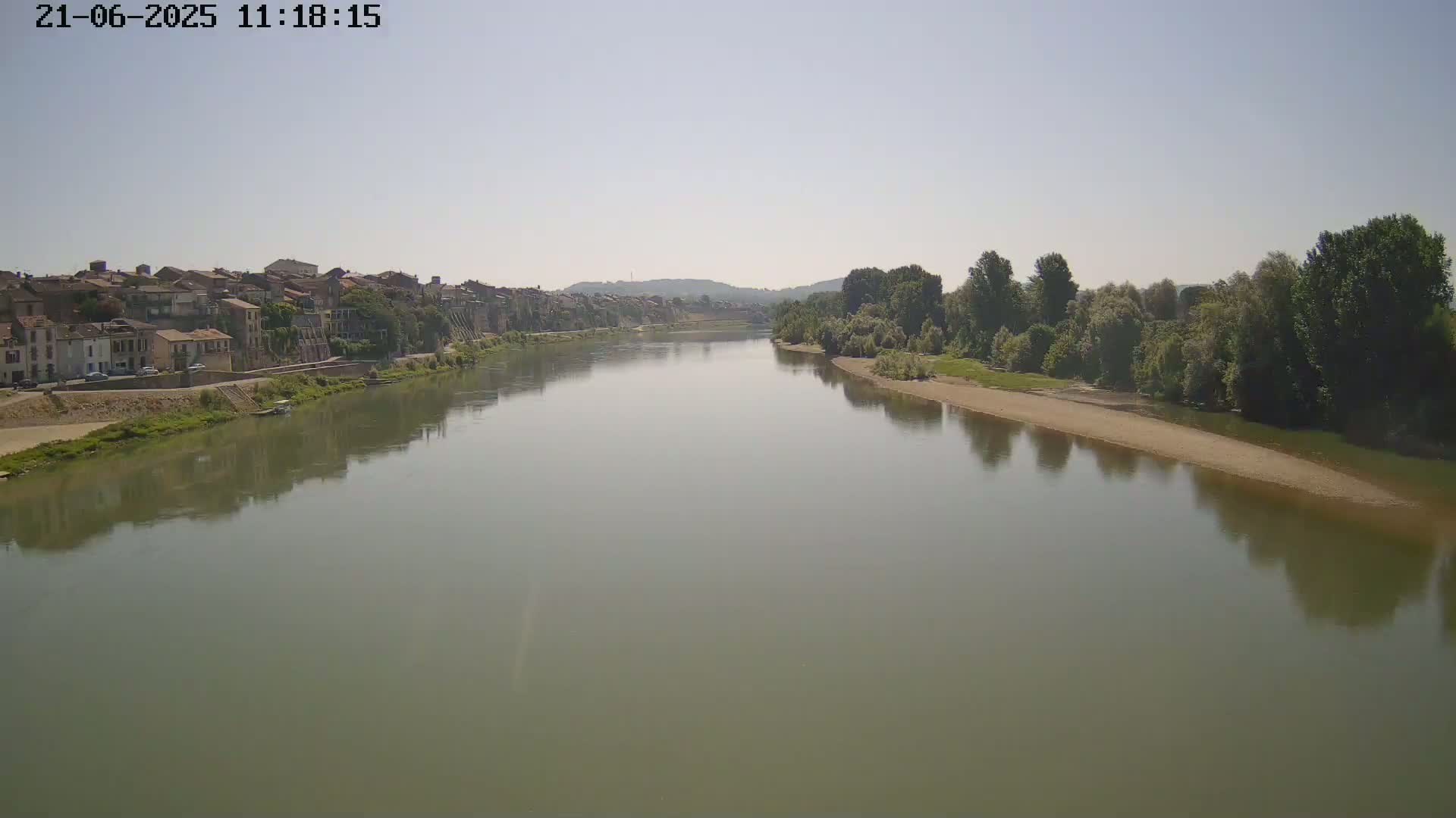 A wide, calm river reflects a town and tree-lined banks under a clear sky.