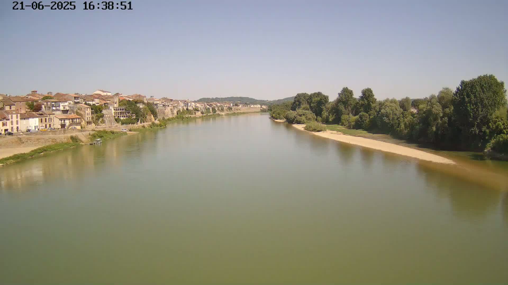 A wide river flows past a town on a clear, sunny day, with trees lining the opposite bank.