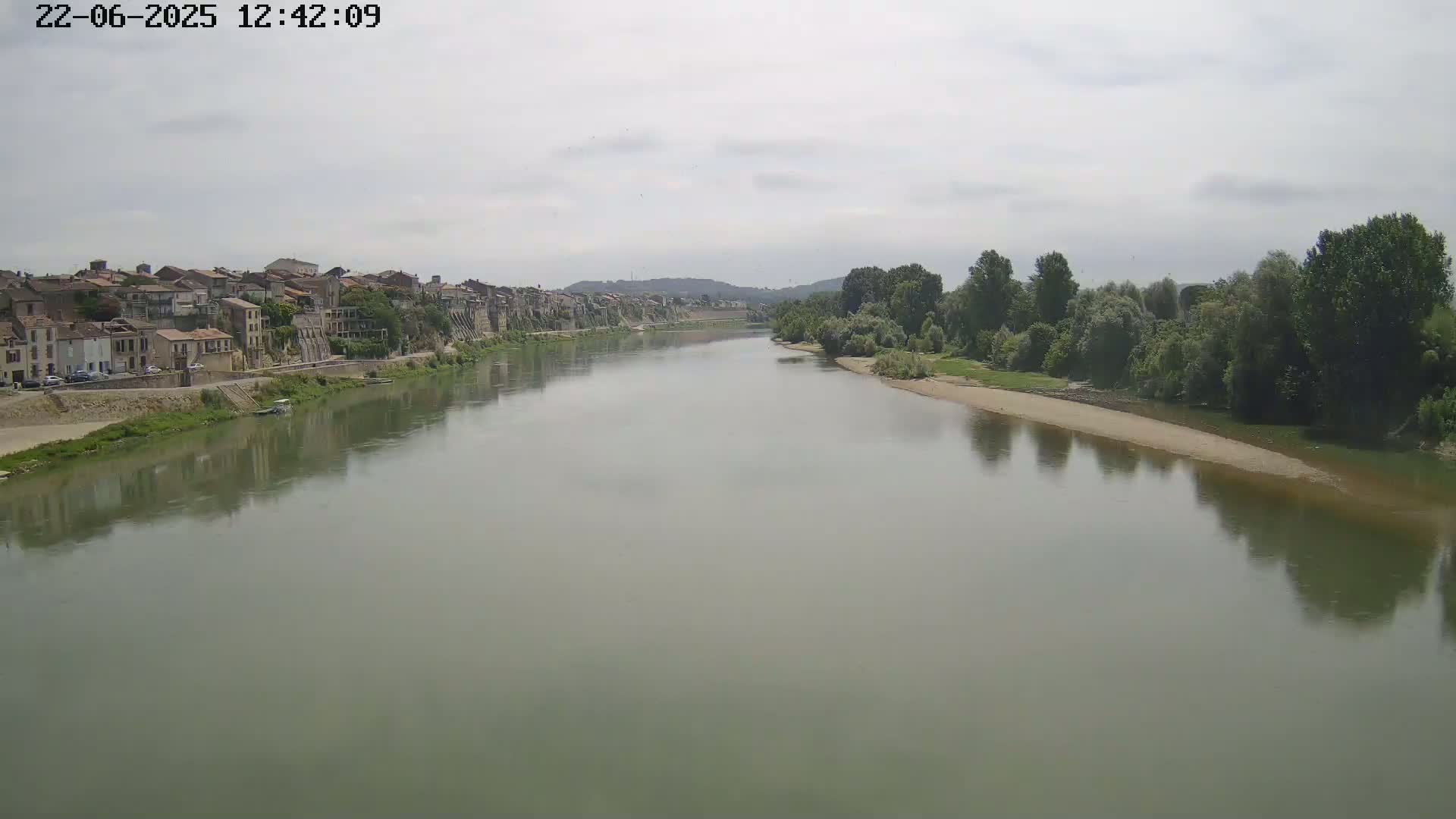 A wide, calm river reflects a cloudy sky and the buildings of a town on its bank, lined with green vegetation on the opposite shore.