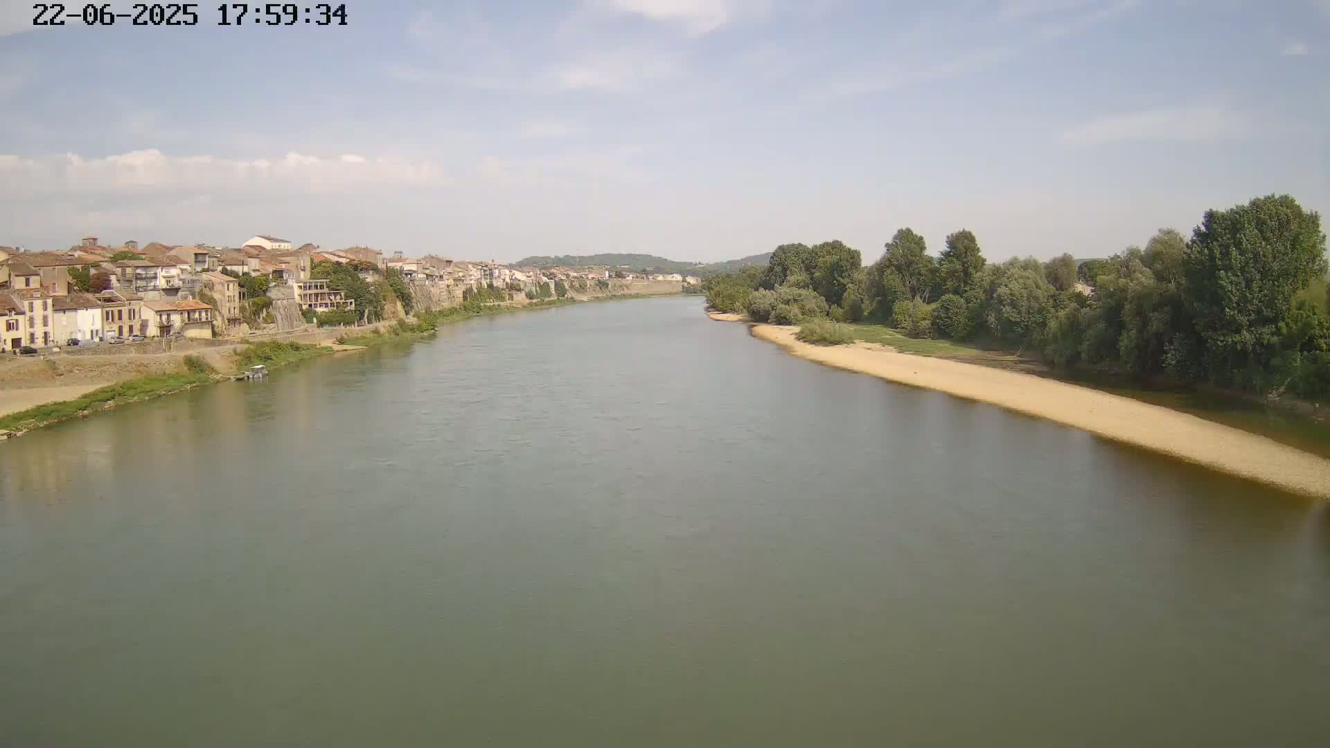 A wide, calm river flows past a town nestled against a hillside under a mostly clear sky.