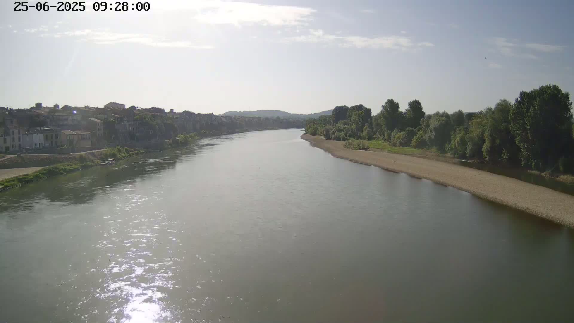 A wide river flows under a mostly sunny sky past a town and a sandy riverbank lined with trees.