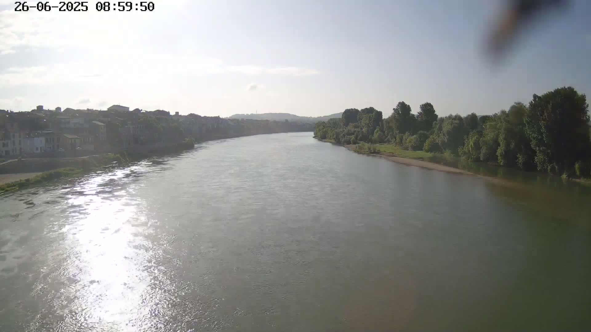 A wide river reflects sunlight on its surface, flowing past a town on one bank and lush green trees on the other under a mostly clear sky.