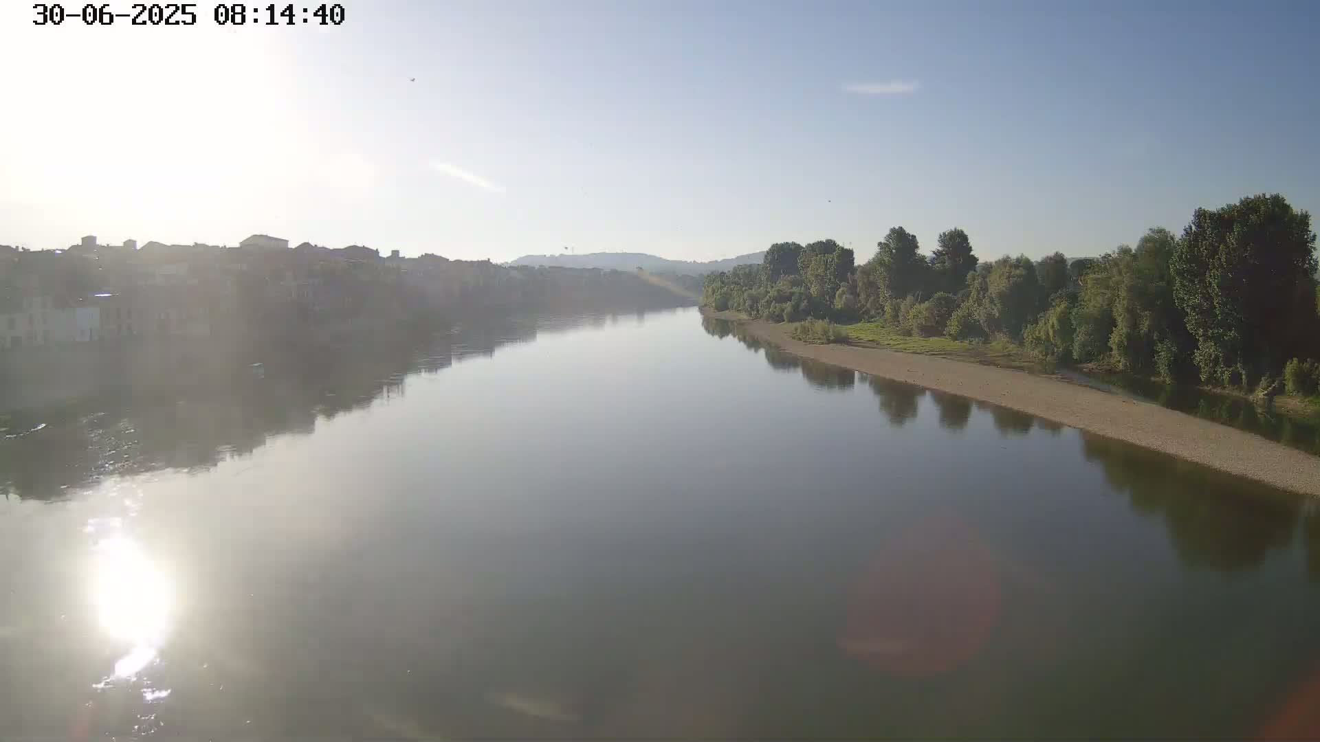 A calm river reflects a hazy sunrise and the buildings and trees on its banks.