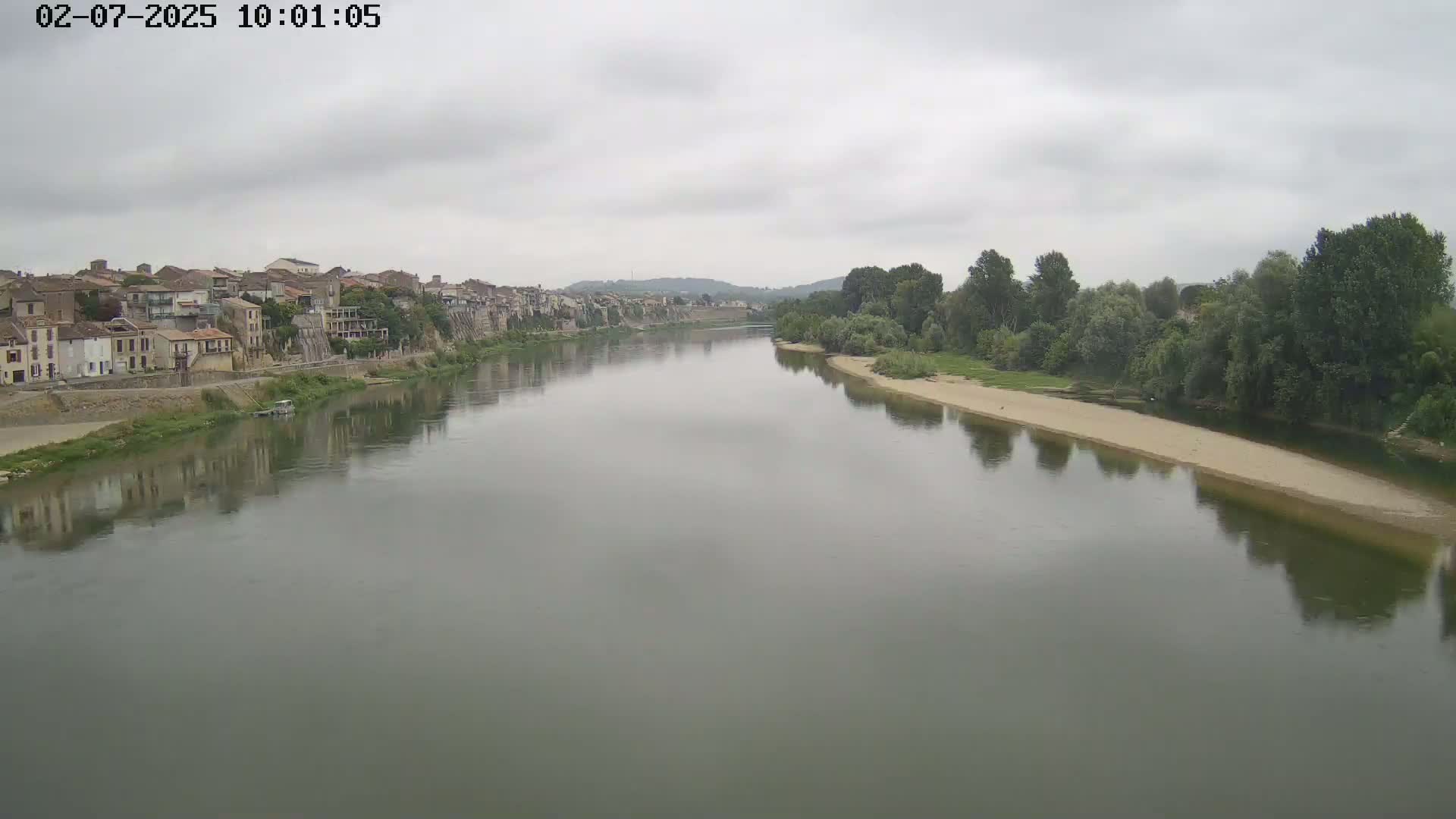 A wide river reflects a cloudy sky and the buildings of a town on its bank, lined with green trees on the opposite side.