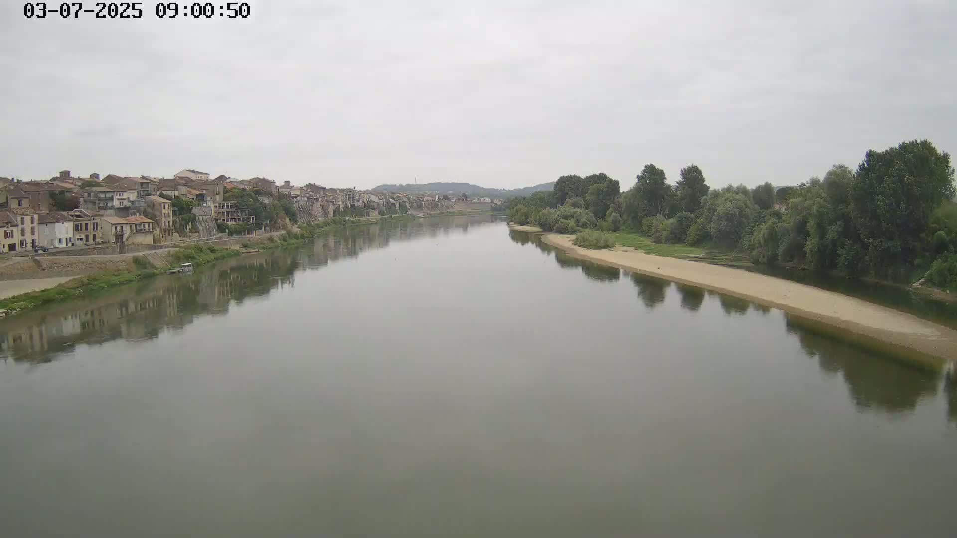 A calm river reflects a town and tree-lined banks under an overcast sky.