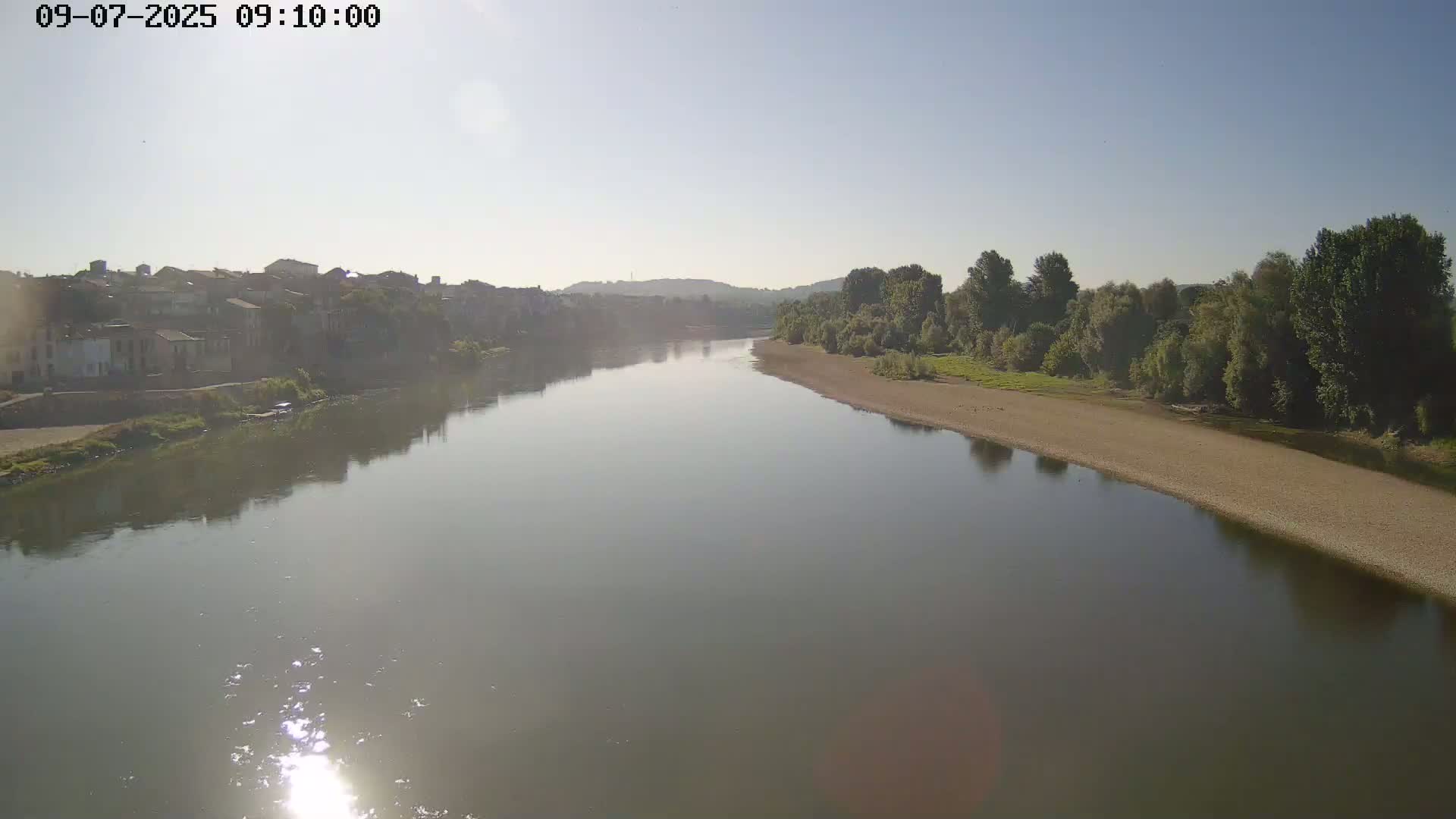A calm river reflects a hazy sunlit sky, flowing past a town and a sandy riverbank lined with green trees.