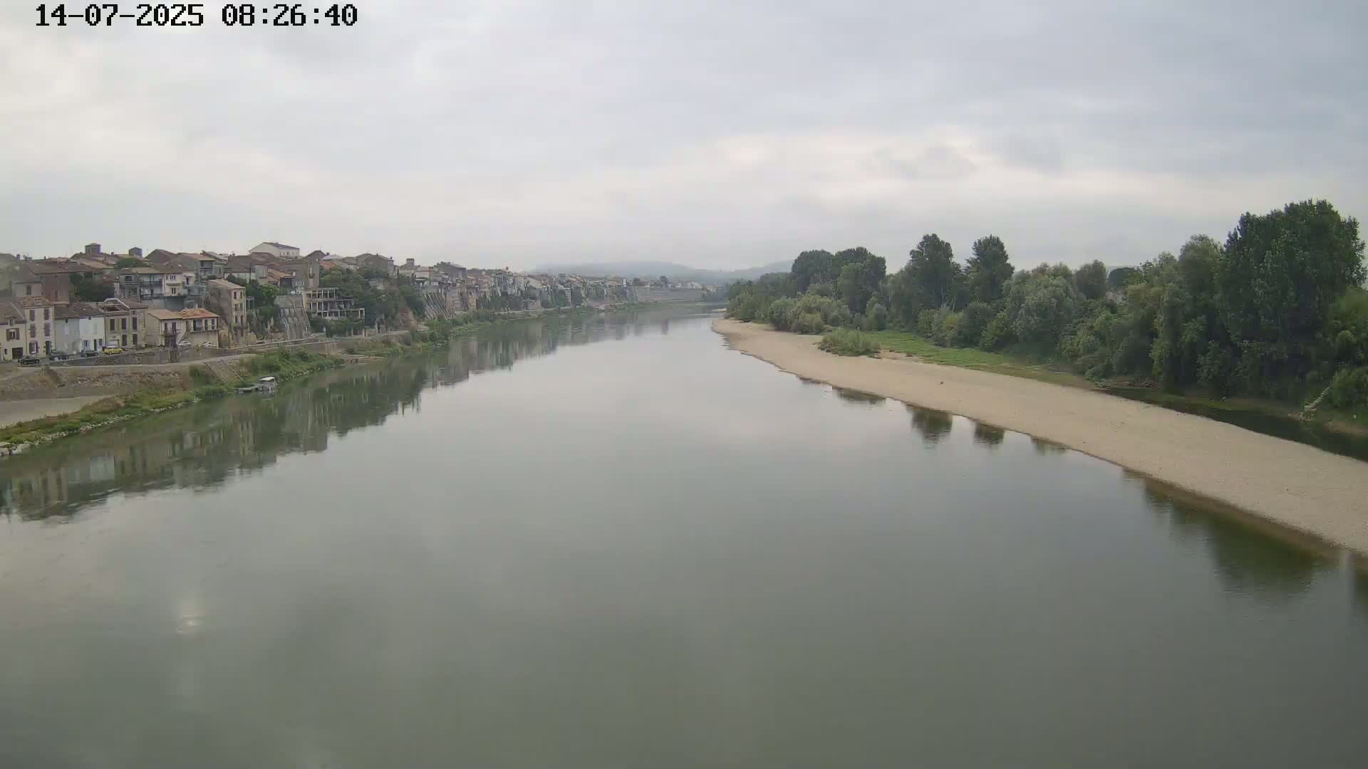 A wide, calm river reflects a town on its bank under an overcast sky.