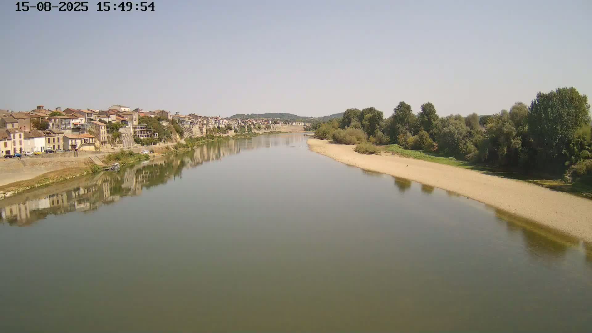 A wide, calm river flows past a town and a sandy riverbank lined with trees under a mostly sunny sky.