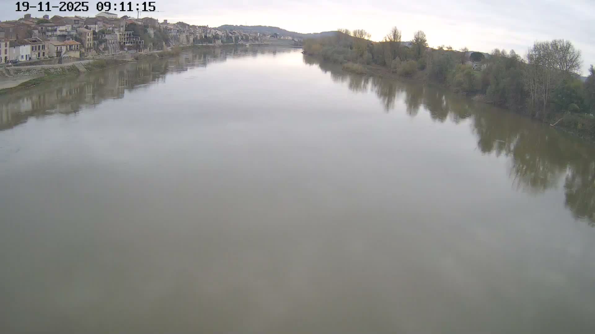 A broad, calm river flows under an overcast sky, bordered by a hillside town with traditional buildings on the left and a dense tree-lined bank on the right, on a cloudy day.