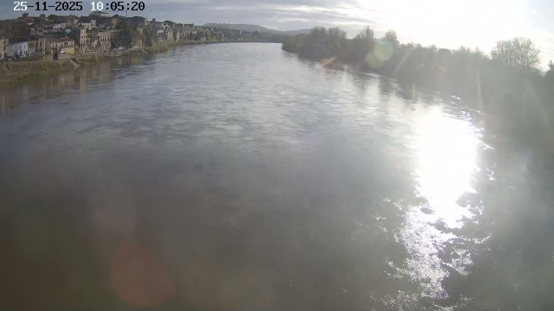 A wide river flows under a partly cloudy, sunny sky with strong sun glare reflecting on its surface, flanked by a town with buildings on the left bank and distant trees on the right bank.