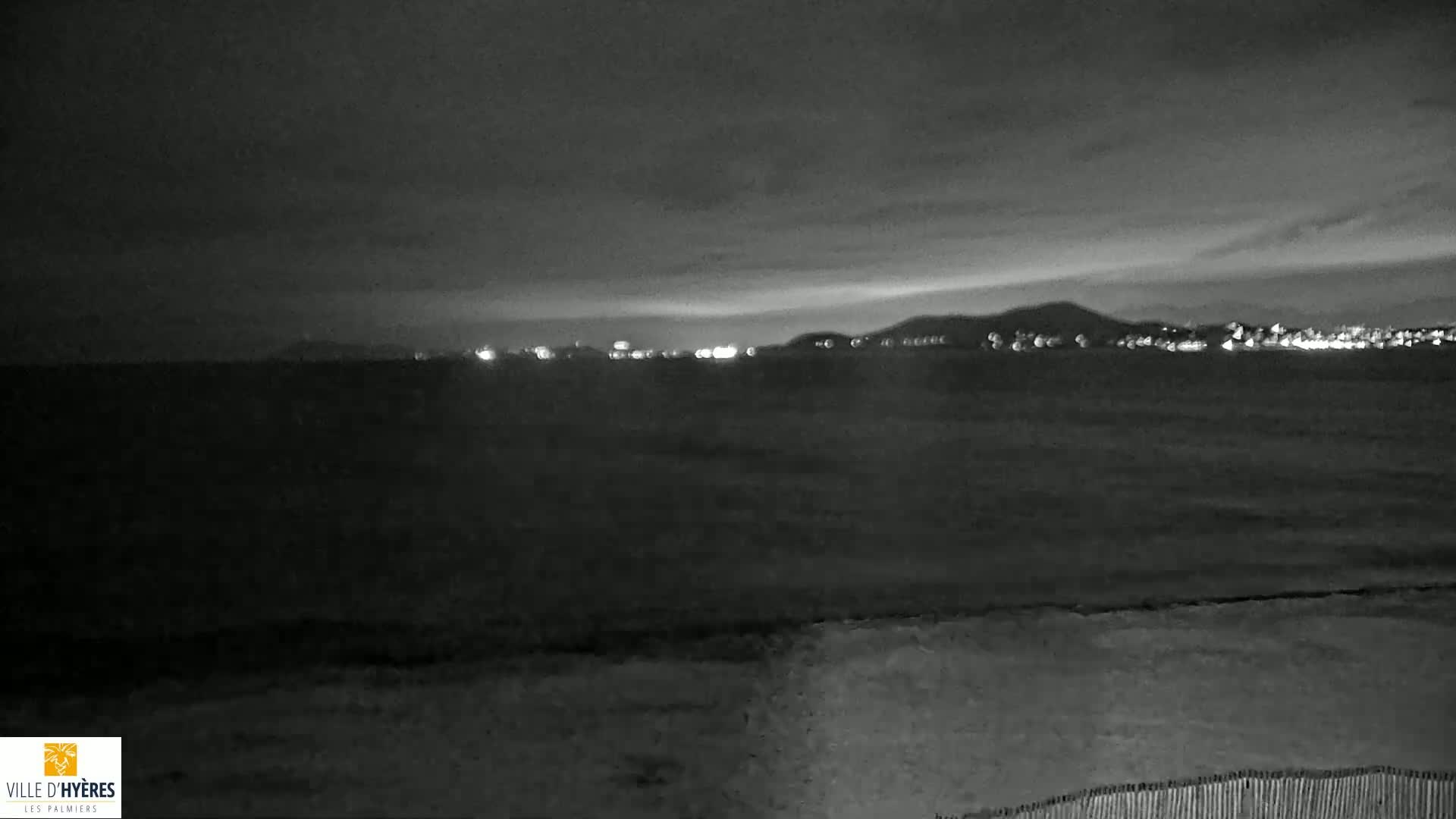 A nighttime view of a dark, calm sea with a distant, faintly lit city skyline under a cloudy sky.