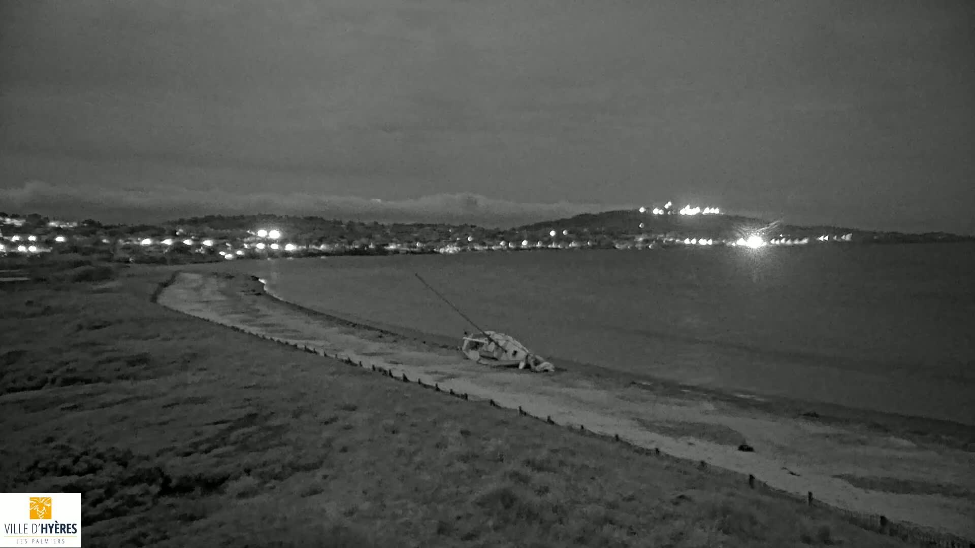 A capsized sailboat lies on a beach at night under a cloudy sky, with a town's lights visible across the water.
