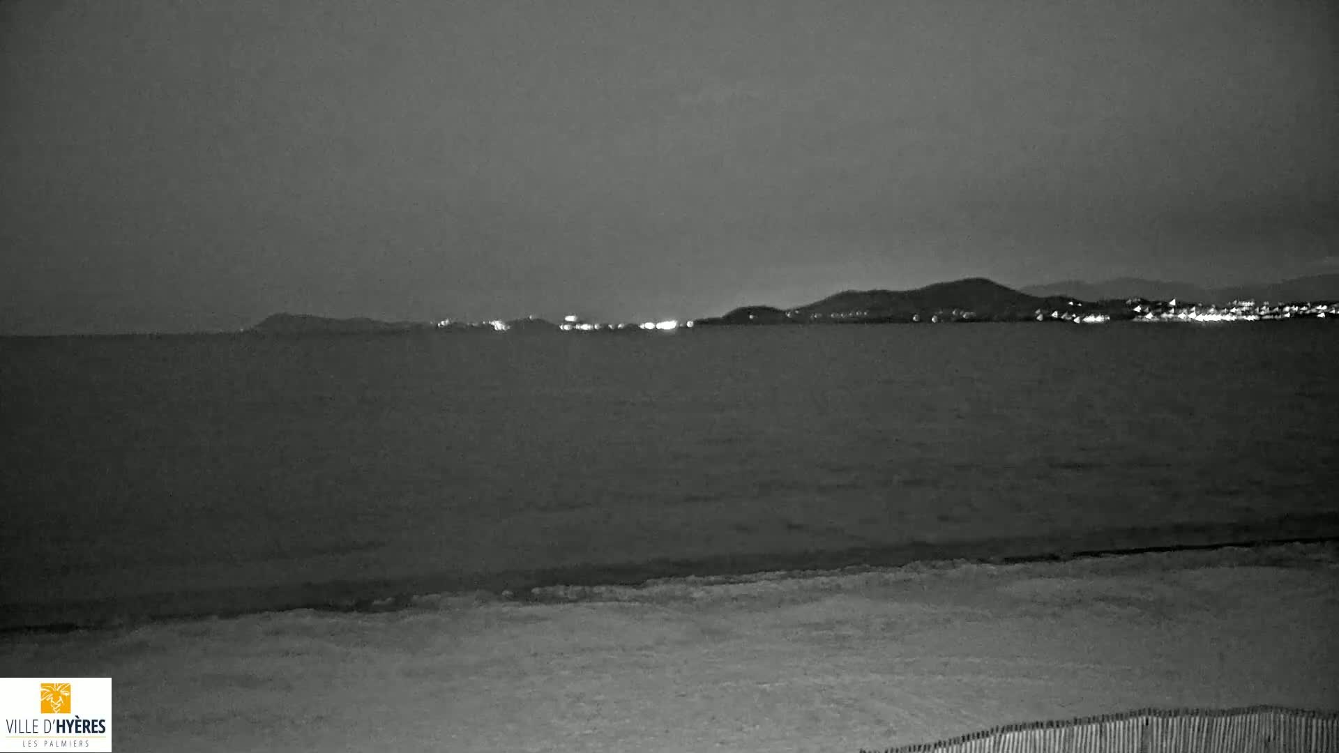 A dark, nighttime beach scene shows a calm sea with a distant, faintly lit shoreline under a cloudy sky.