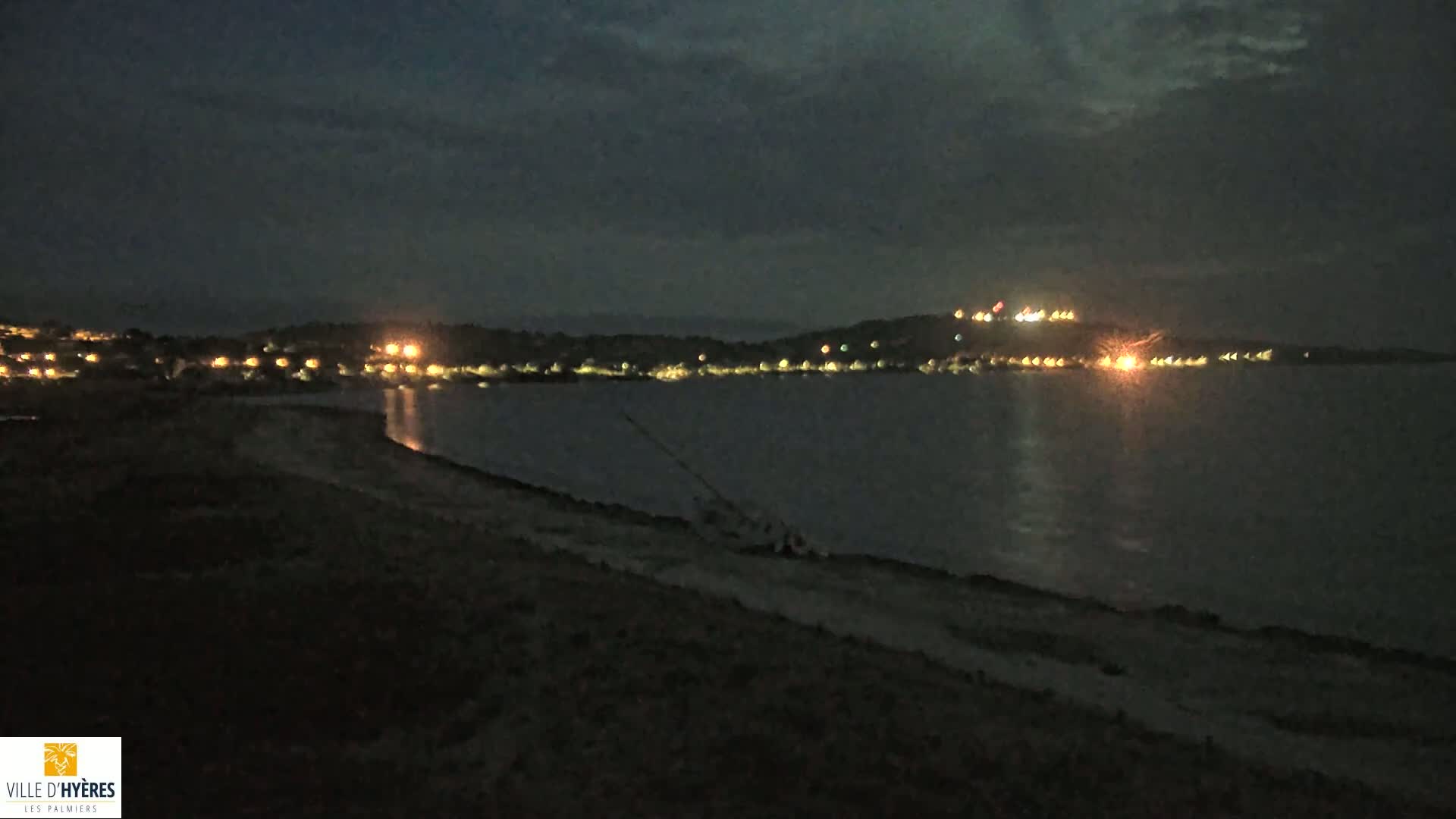 A dark, nighttime scene shows a body of water reflecting lights from a distant town on the shore, with a beached sailboat visible in the foreground under a cloudy sky.