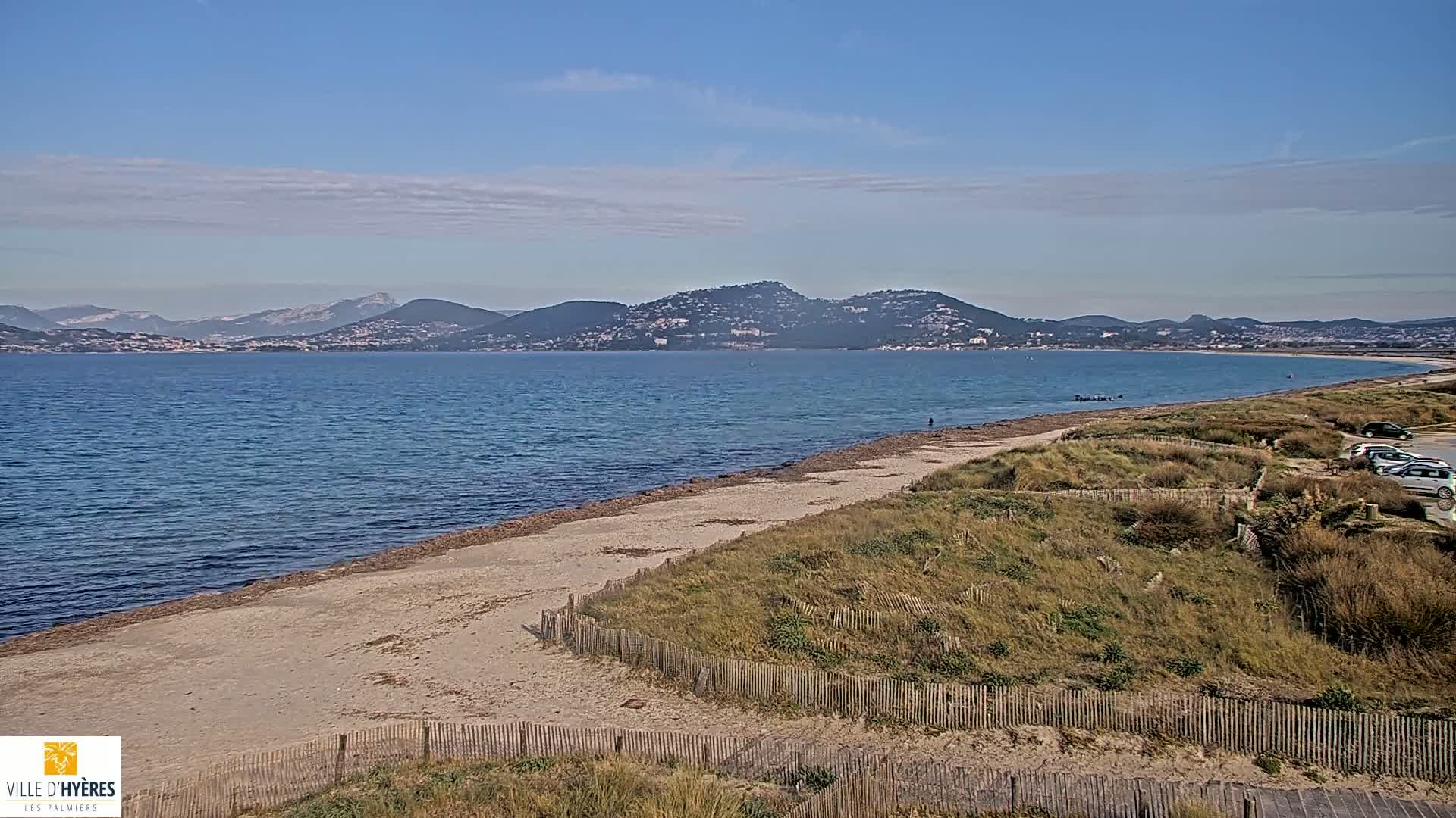 A sandy beach stretches along a calm, blue body of water, with mountains visible in the distance under a mostly clear sky.