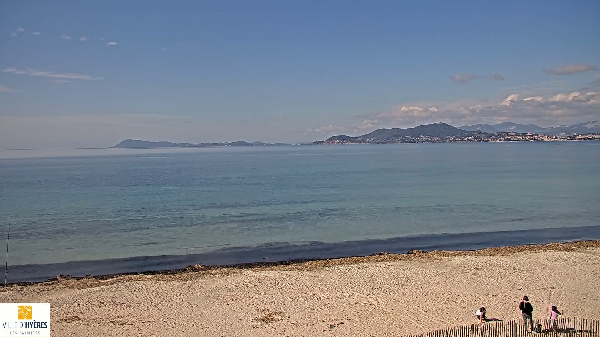 A sandy beach stretches towards a calm, light-blue sea under a mostly clear sky, with distant hills and islands visible on the horizon.