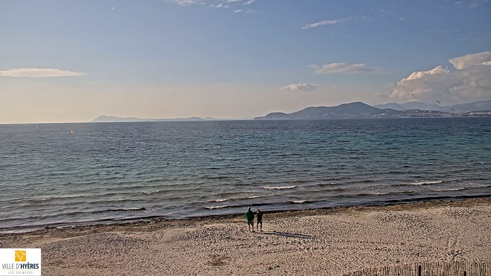 Two people stand on a sandy beach overlooking a calm sea with distant mountains under a partly cloudy sky.