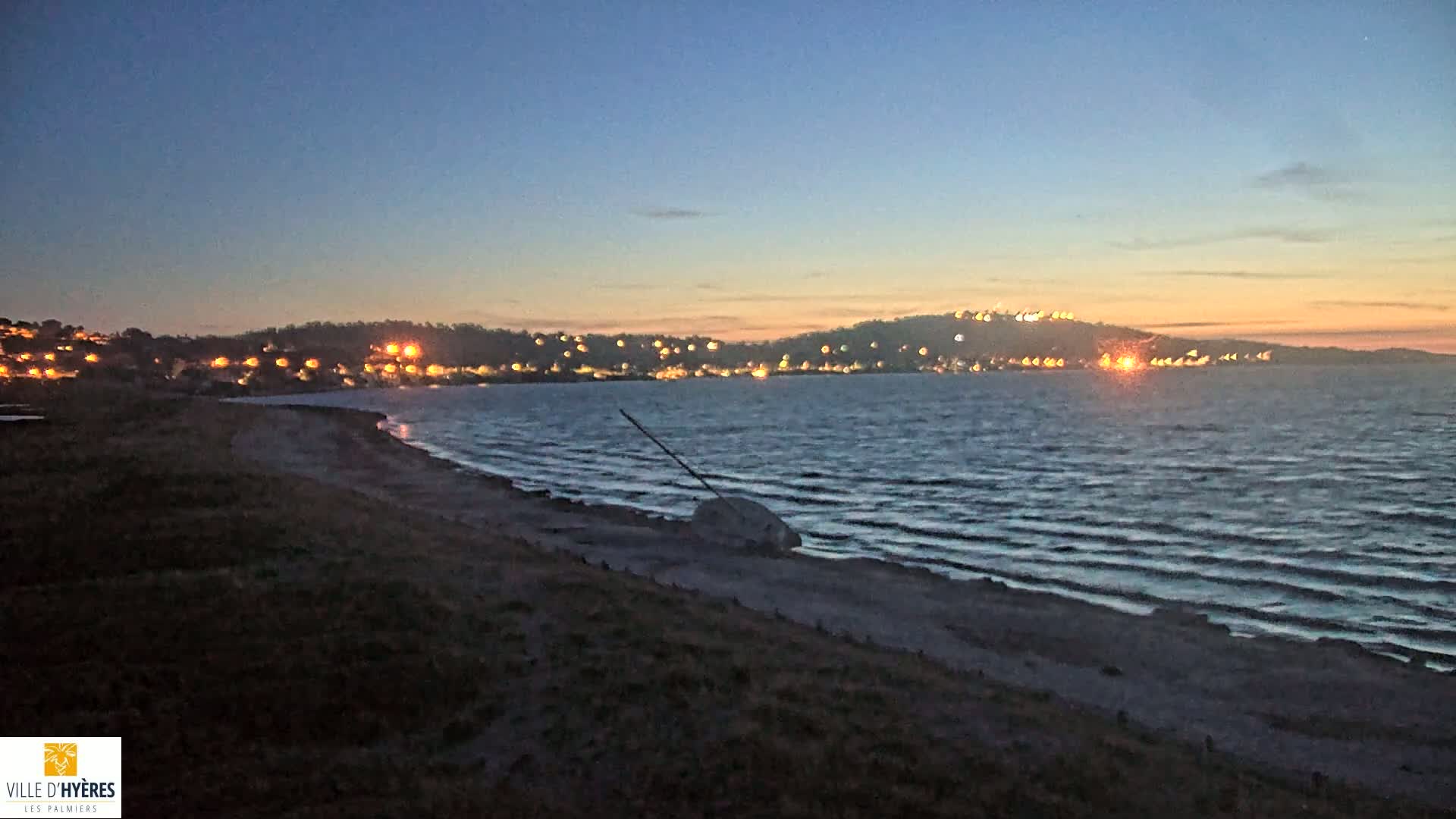 A dark beach at twilight shows a partially submerged object on the shore, with a town's lights illuminating the opposite side of a calm, dark-blue ocean.