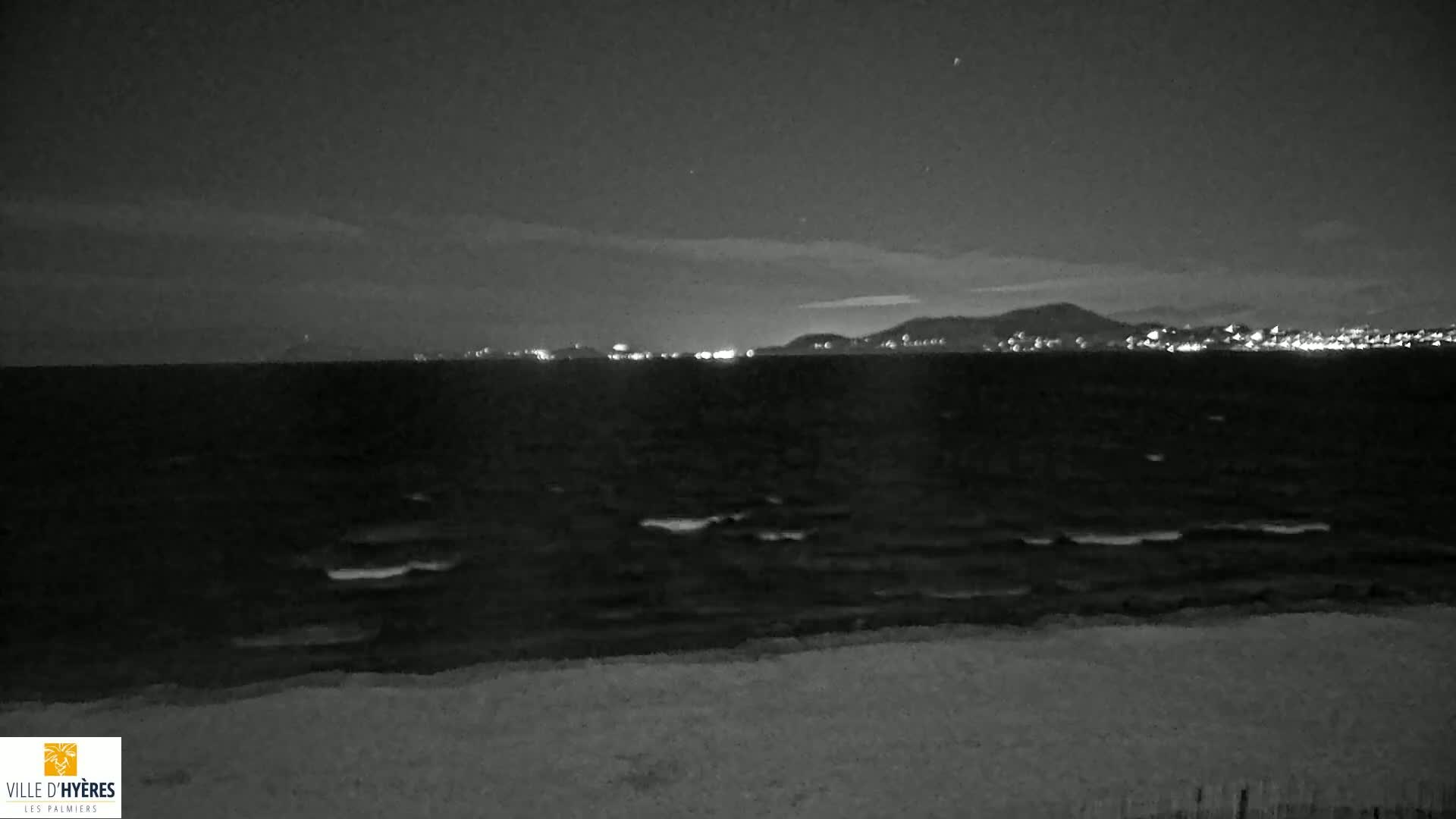 A dark, nighttime beach scene shows a gently waving sea under a partly cloudy sky with city lights visible in the distance.