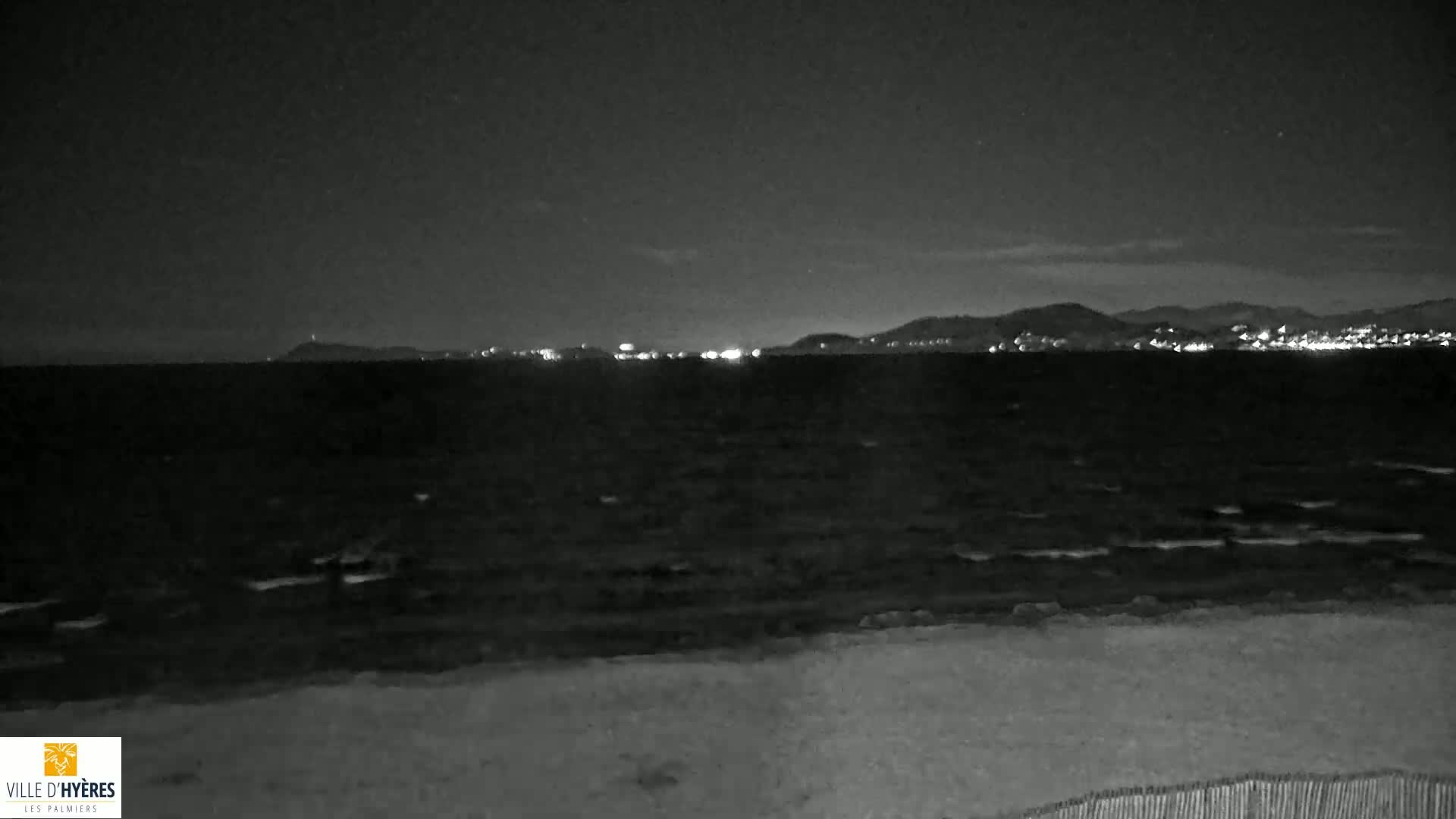 A nighttime view of a dark, wavy ocean with a distant, faintly lit cityscape and hills under a mostly clear sky.