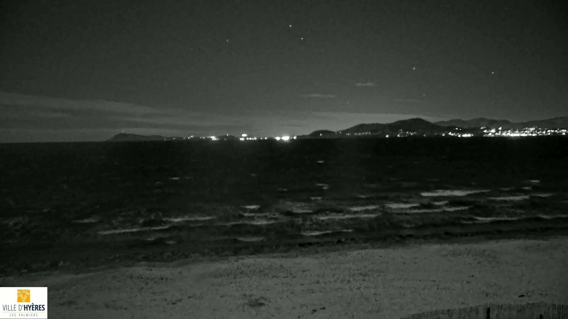 A dark, nighttime beach scene shows a gently rolling ocean under a slightly cloudy sky with a distant city skyline faintly illuminated.