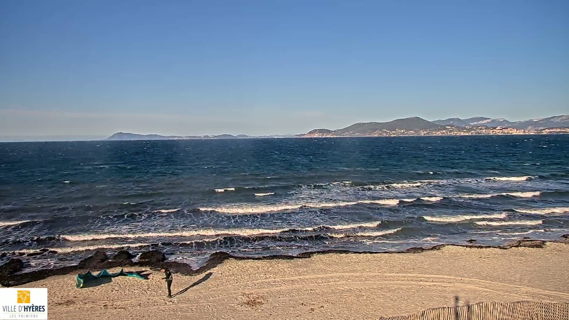 A person stands on a sandy beach near a dark-colored shoreline, looking out at a wavy ocean under a clear blue sky, with distant mountains visible on the horizon.