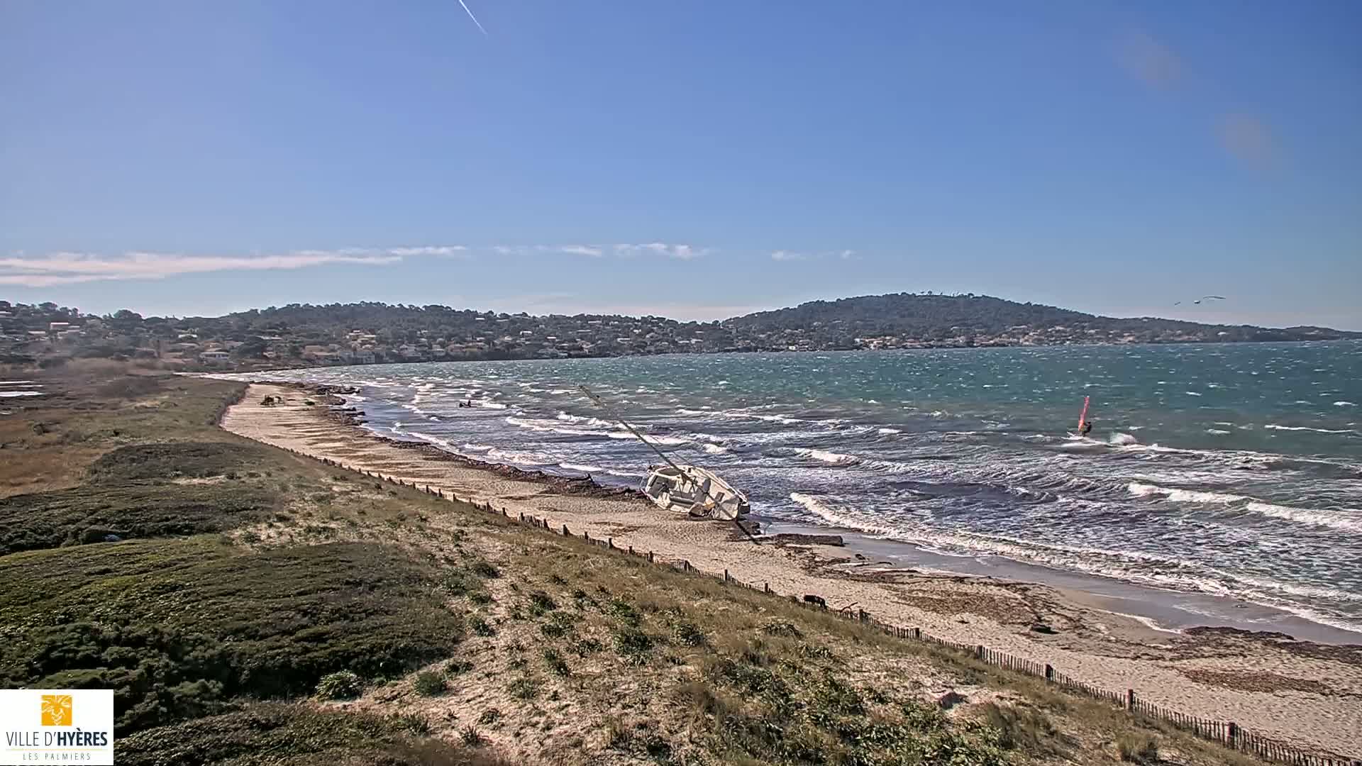 A sandy beach with a capsized sailboat on the shore, under a mostly sunny sky with moderate waves crashing on the beach and a windsurfer in the water.