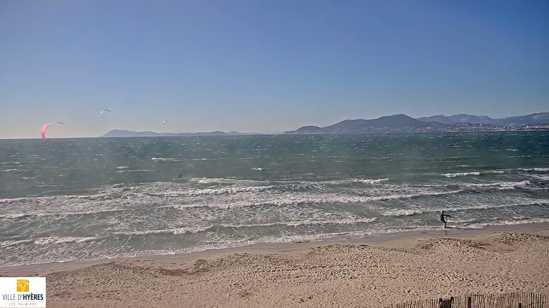 A sandy beach with several kitesurfers in the water under a clear blue sky, with a windy sea and distant mountains visible on the horizon.