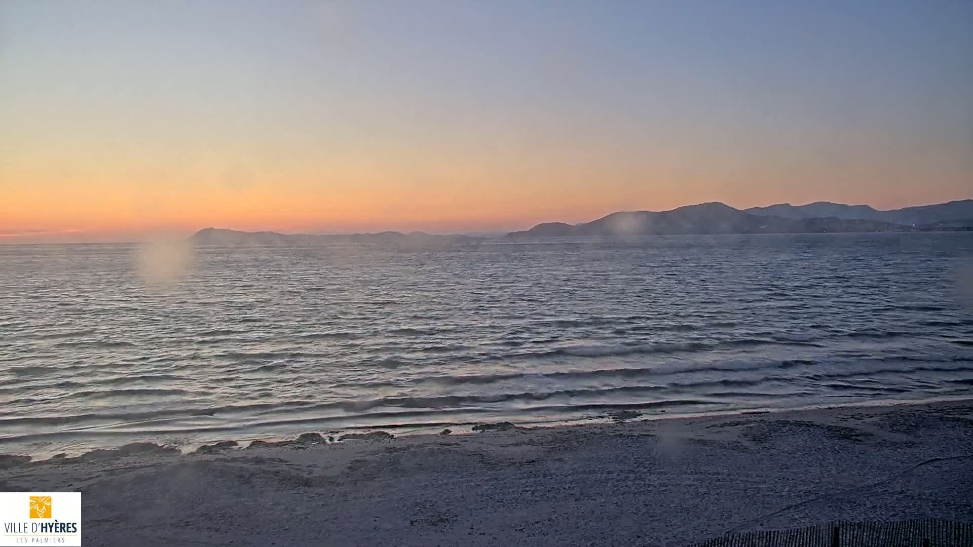 A sandy beach at sunset overlooks a calm sea with small waves, and distant mountains under a hazy, pinkish sky.
