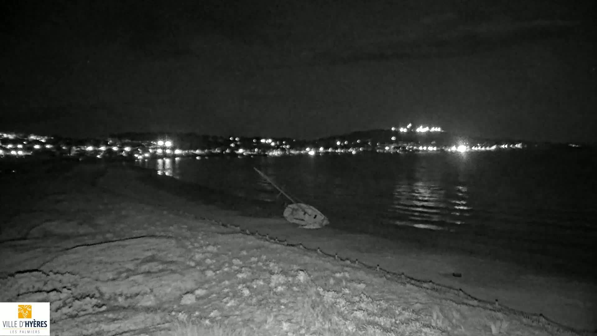A small boat lies beached on a sandy shore at night, with a town's lights visible across the calm water.