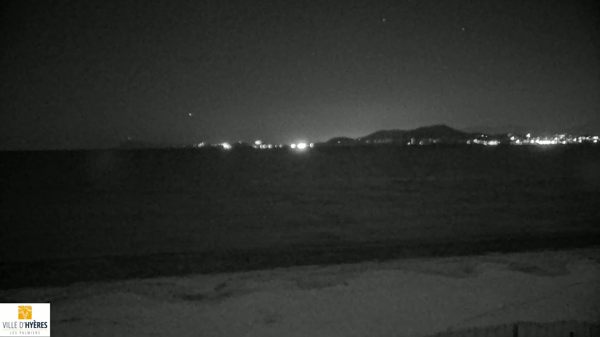 A dark, nighttime beach scene shows a calm body of water with a distant city skyline illuminated under a clear, star-studded sky.