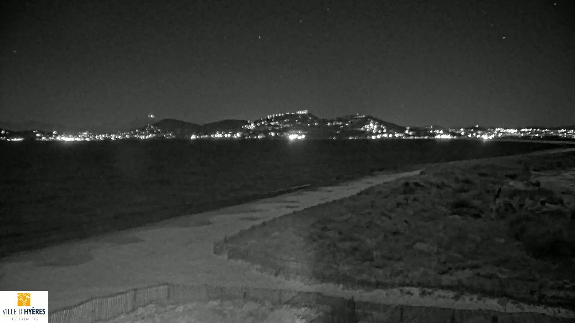 A nighttime view of a city's lights across a body of water, from a dark, sandy shore.