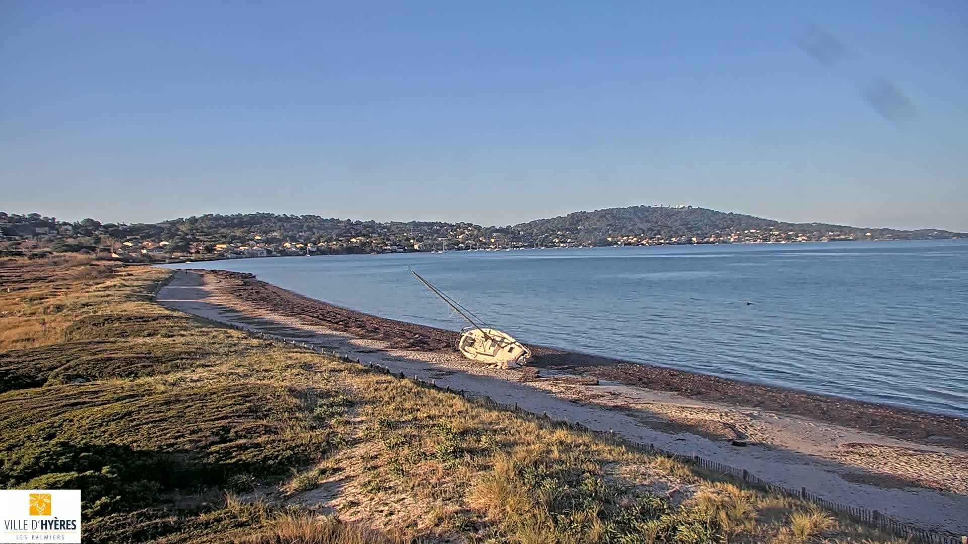 A capsized sailboat lies on a sandy beach next to calm blue water under a clear blue sky, with a town visible in the distance.