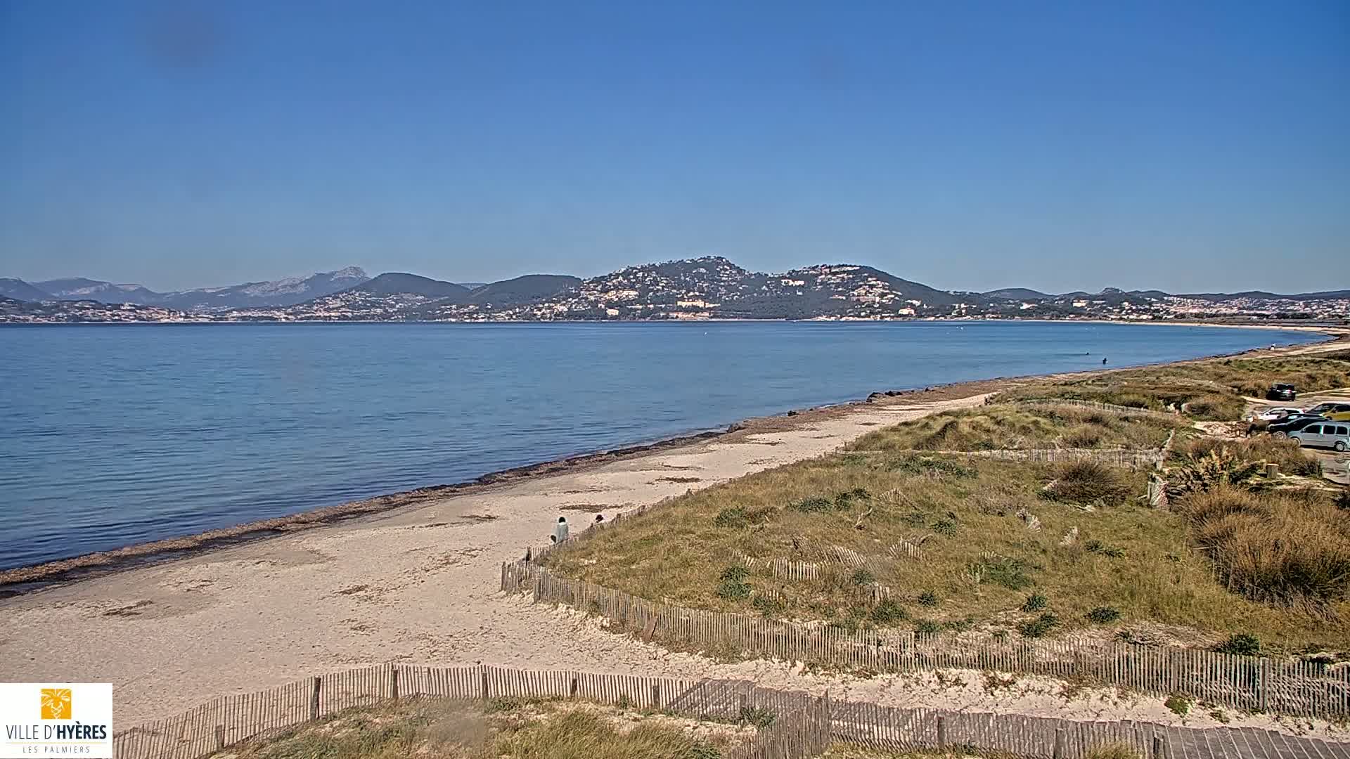 A sandy beach with a wooden fence and a few people stretches along a calm, blue sea under a clear sky, with mountains visible in the distance.