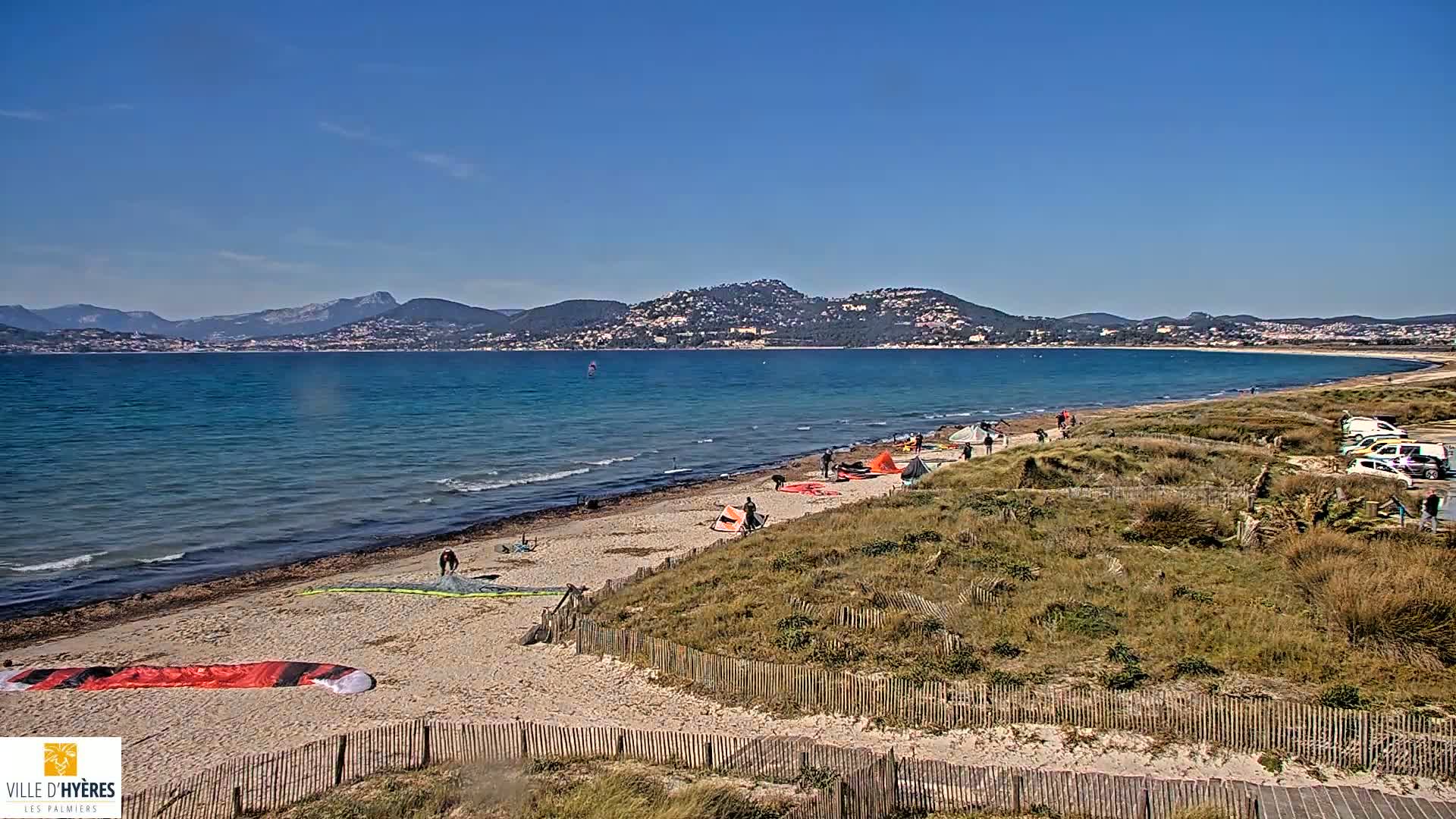 A sandy beach with kite surfers and several kites on the sand, overlooks a calm blue ocean and distant mountains under a clear blue sky.