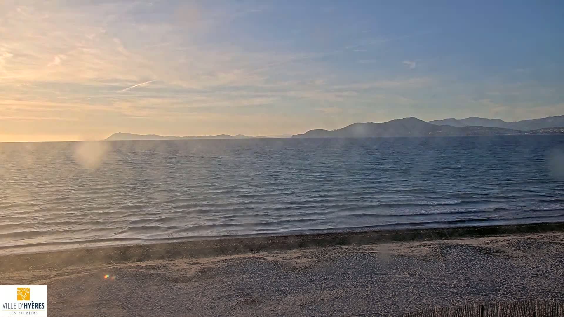 A sandy beach meets a gently wavy sea under a hazy, pale sunset sky with distant mountains visible on the horizon.