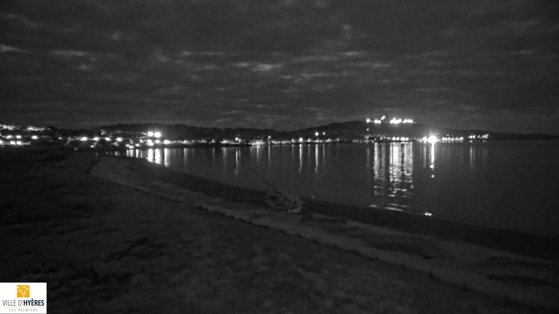 A nighttime, black and white view of a calm body of water reflecting the lights of a distant town under a cloudy sky, with a dark shoreline in the foreground containing some debris.