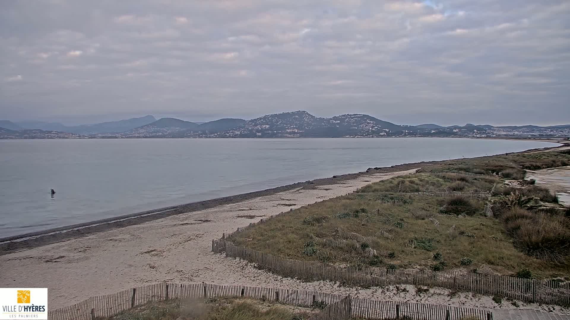 A sandy beach stretches along a calm, gray body of water under an overcast sky, with distant mountains visible on the horizon.